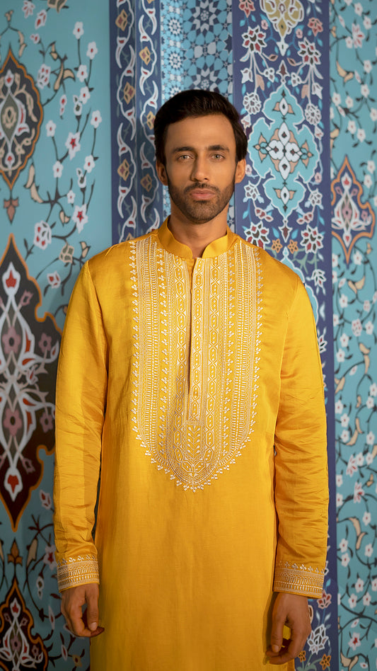Man wearing a mustard embroidered kurta against a decorative blue wall.
