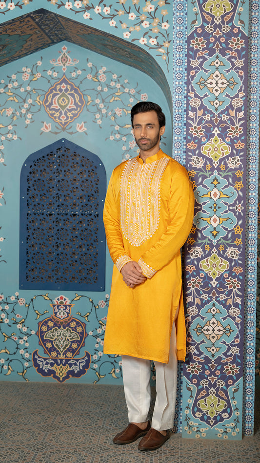 Man in a mustard kurta standing against a decorative wall.