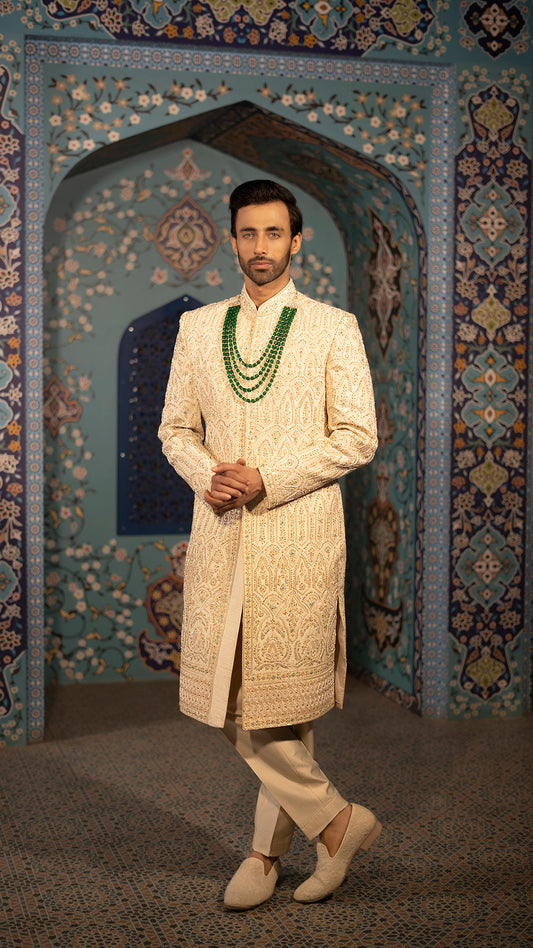 Man wearing a traditional cream sherwani with intricate patterns in the background