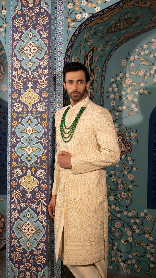 Man in a beige sherwani with intricate patterns against a decorative wall.