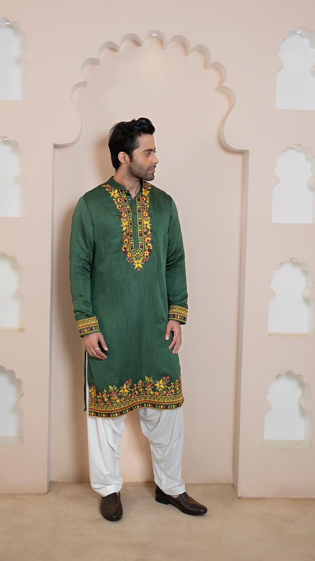 Man wearing a green embroidered kurta with white pants against a decorative wall.
