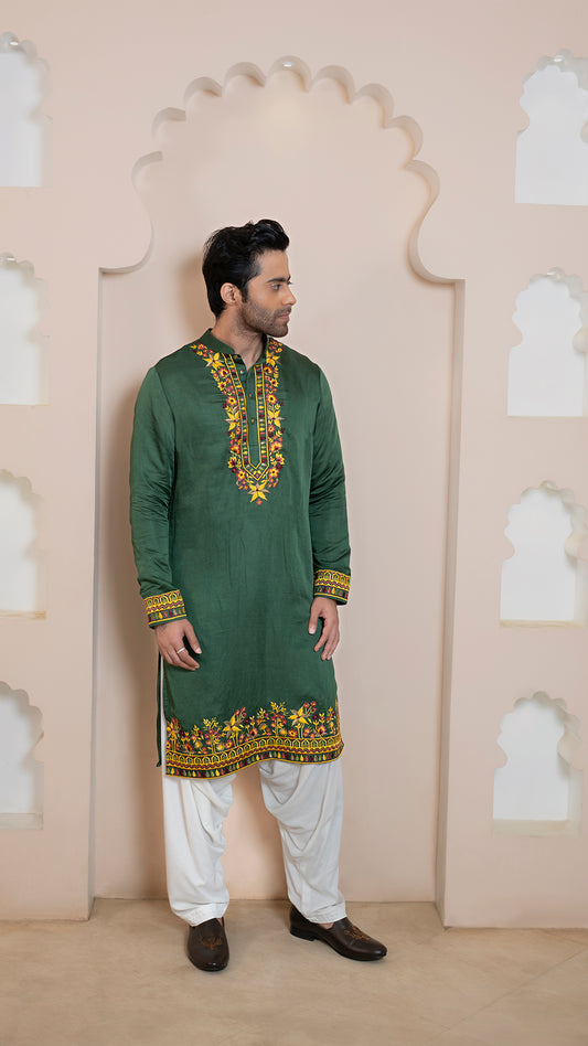 Man wearing a green embroidered kurta with white pants against a decorative wall.