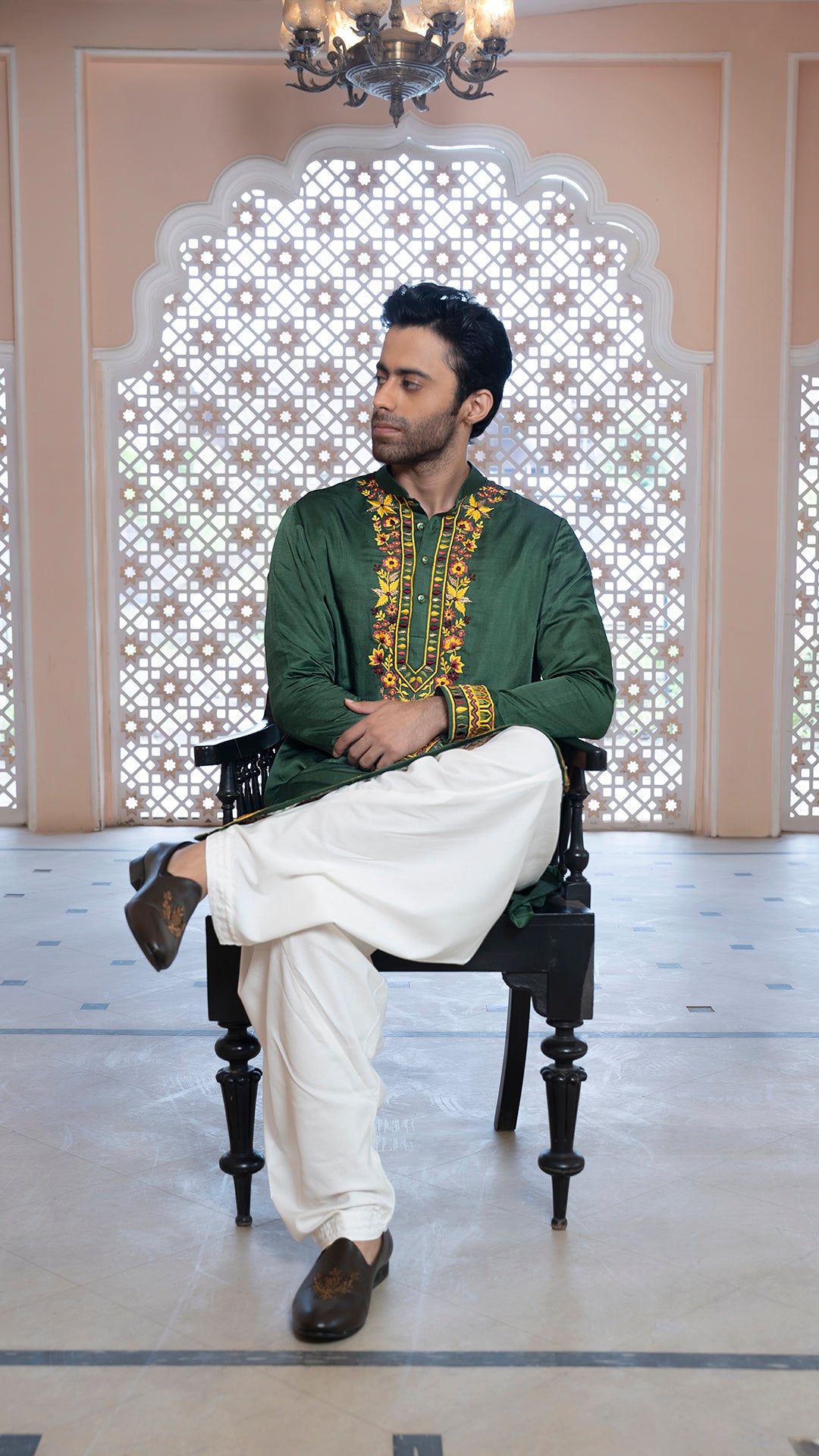 Man sitting on a chair wearing a green traditional outfit with white pants in a decorated room.