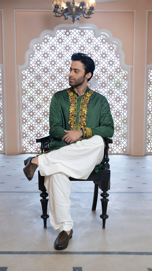 Man sitting on a chair wearing a green traditional outfit with white pants in a decorated room.
