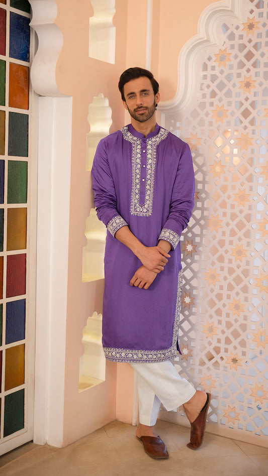 Man wearing a purple embroidered kurta with white pants in a decorative interior setting.