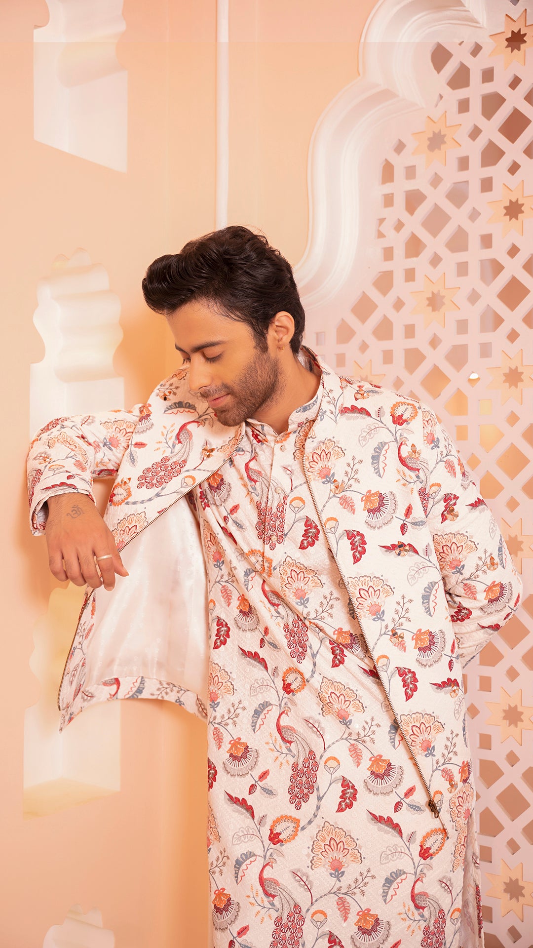 Man wearing a floral patterned kurta jacket against an ornate wall.