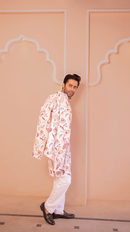 Man wearing a patterned kurta and white pants against a beige wall.