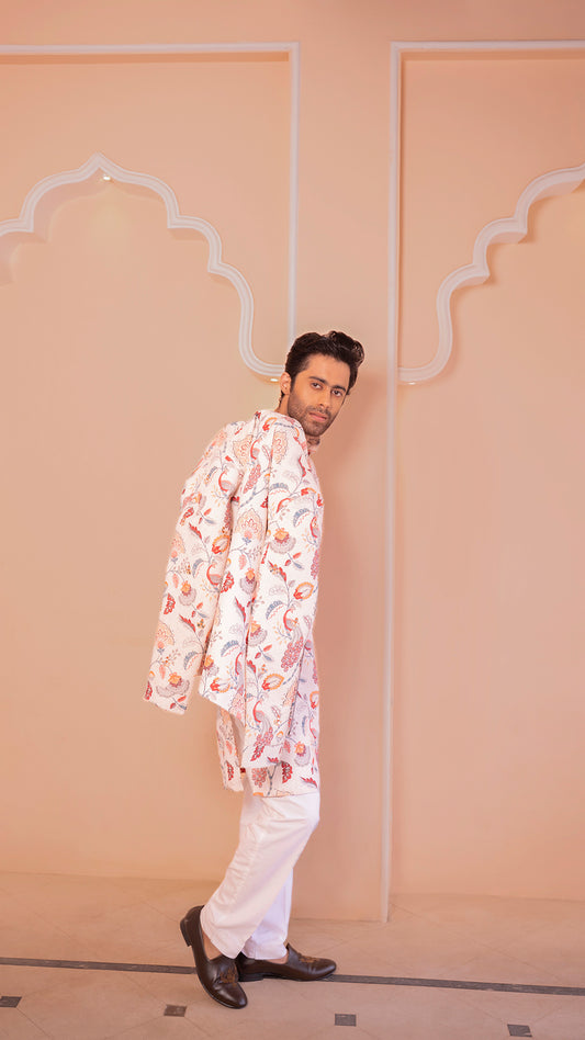 Man wearing a patterned kurta and white pants against a beige wall.
