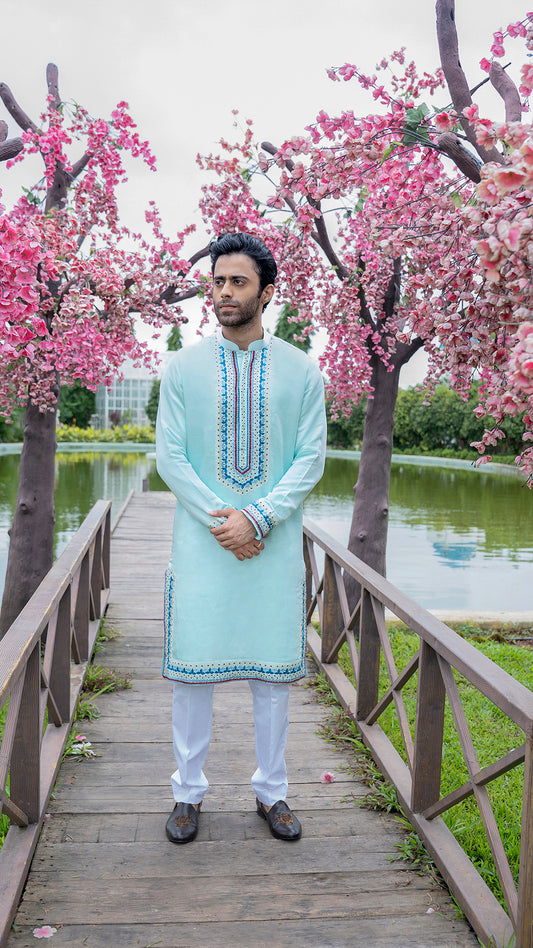 Man in a mint blue kurta with white pants standing on a wooden bridge with cherry blossom trees in the background.