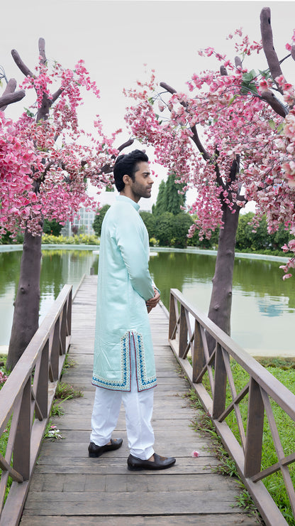Man in a light blue kurta standing on a wooden bridge with cherry blossom trees and a pond in the background.