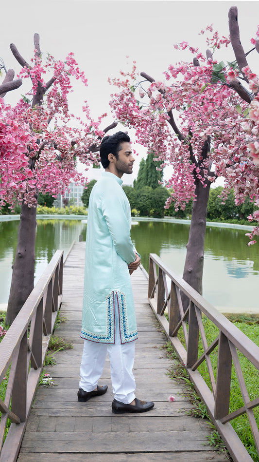 Man in a light blue kurta standing on a wooden bridge with cherry blossom trees and a pond in the background.