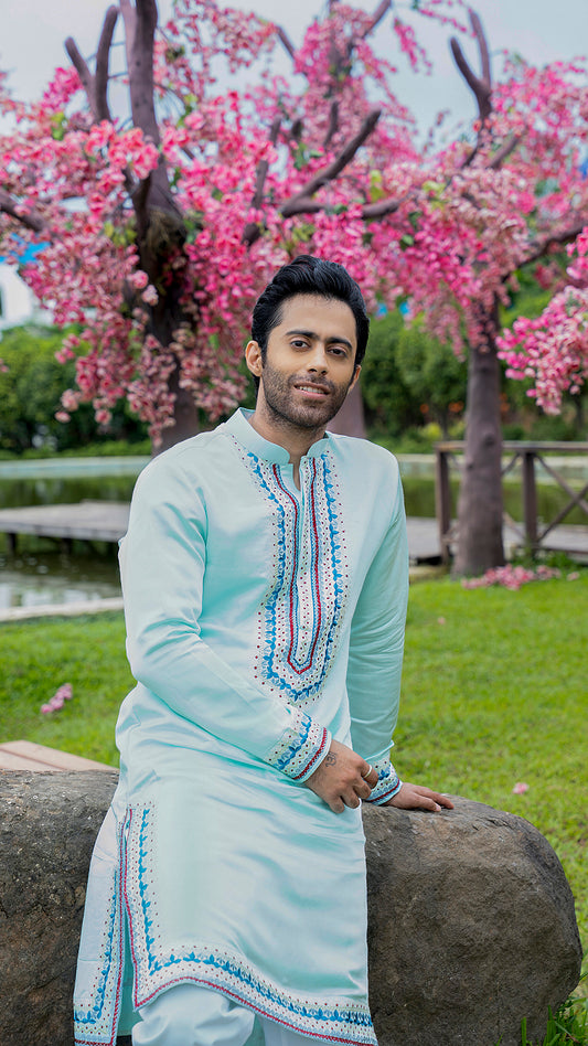 Man in a light blue kurta with embroidery sitting on a rock with pink flowering trees in the background.