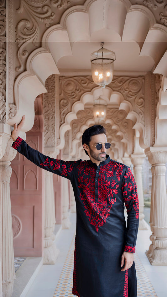 Man in a black and red embroidered traditional outfit standing in an ornate architectural setting.
