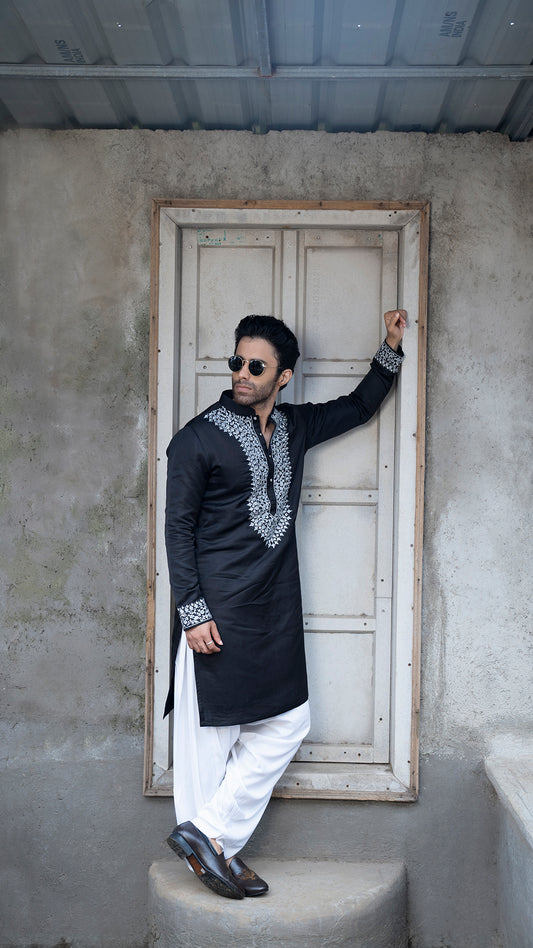 Man in traditional black and grey outfit standing against a plain wall.