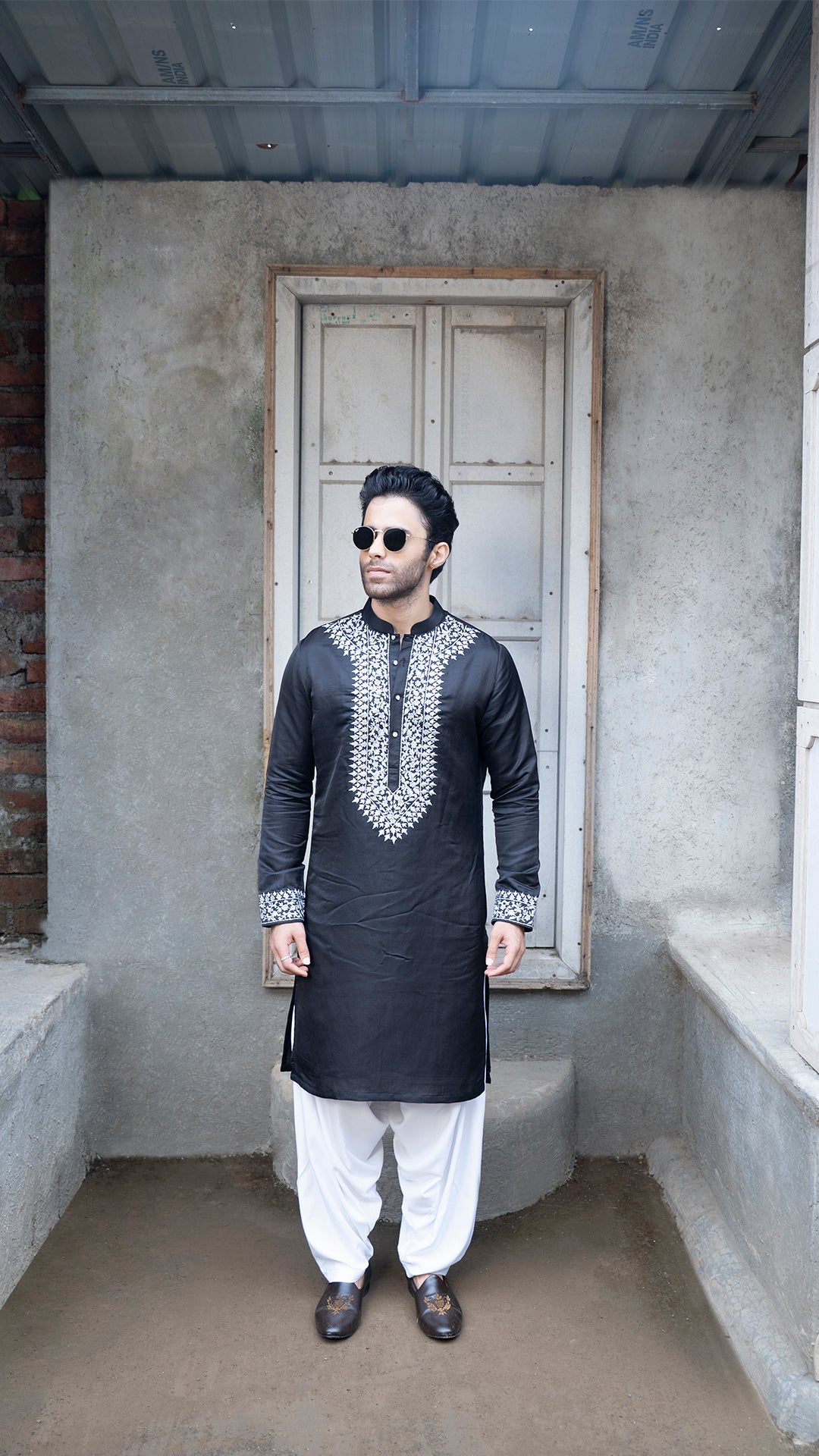 Man wearing a black kurta with grey embroidery standing in a room with concrete walls and a door.