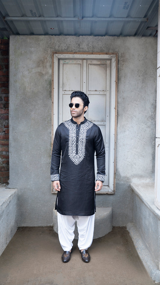Man wearing a black kurta with grey embroidery standing in a room with concrete walls and a door.