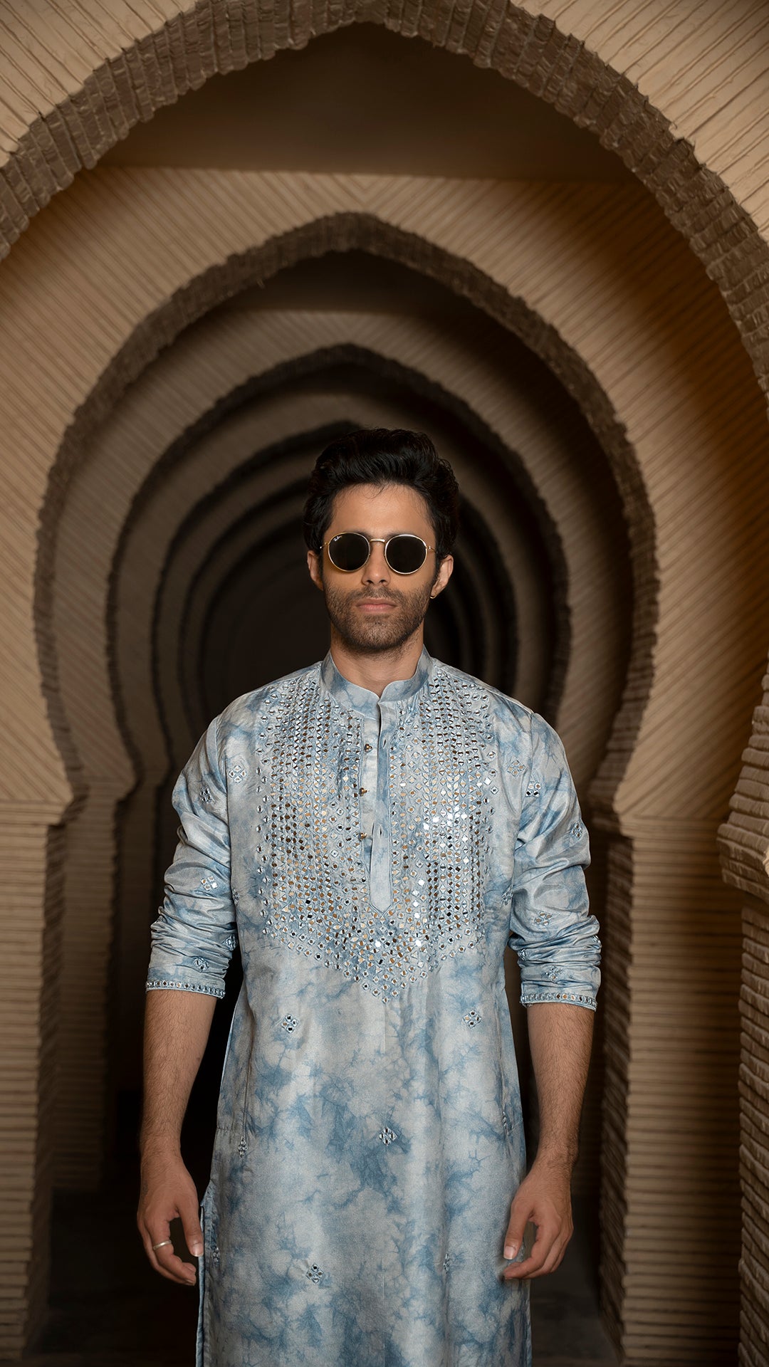 Man wearing a tye-dye blue embroidered kurta standing in front of a series of arches.