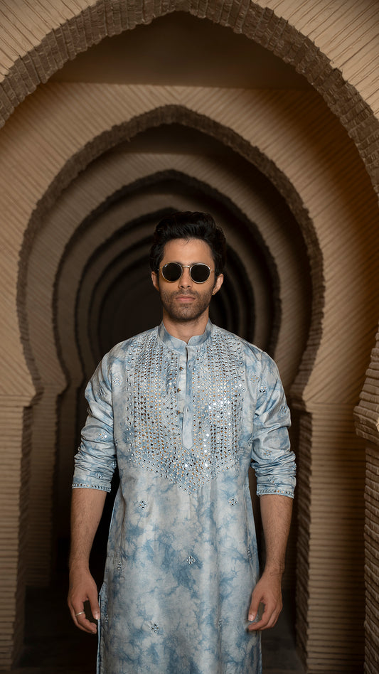 Man wearing a tye-dye blue embroidered kurta standing in front of a series of arches.