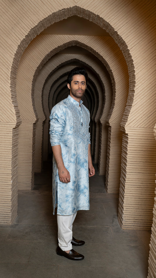 Man in a blue tye-dye kurta standing in a architectural archway.