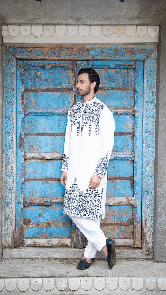 Man wearing a white kurta with blue embroidery standing in front of a blue wooden door.