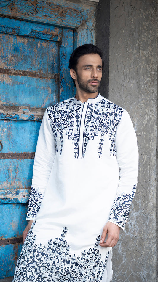 Man wearing a white kurta with blue patterns standing against a blue door and gray wall.