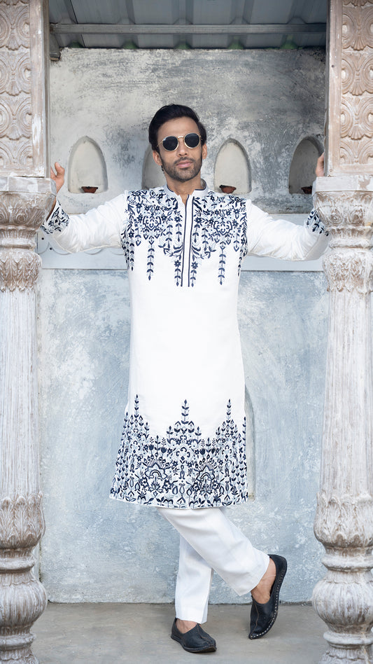 Man wearing a white kurta with blue embroidery standing in front of stone arches.
