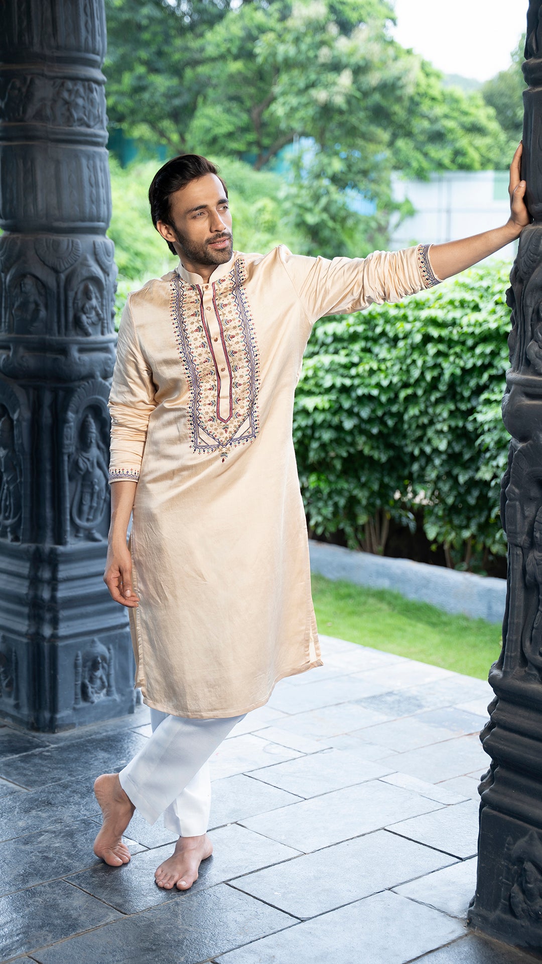 Man in a beige kurta with white pants standing outdoors near decorative pillars.