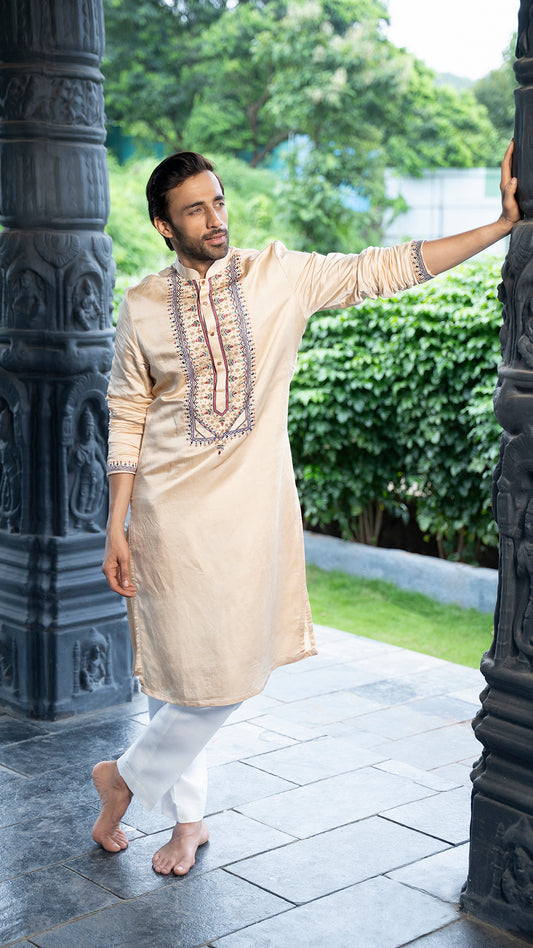 Man in a beige kurta with white pants standing outdoors near decorative pillars.