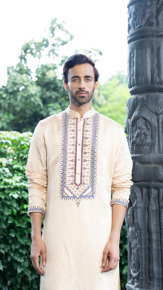 Man wearing a cream kurta with intricate embroidery standing outdoors next to a stone column.
