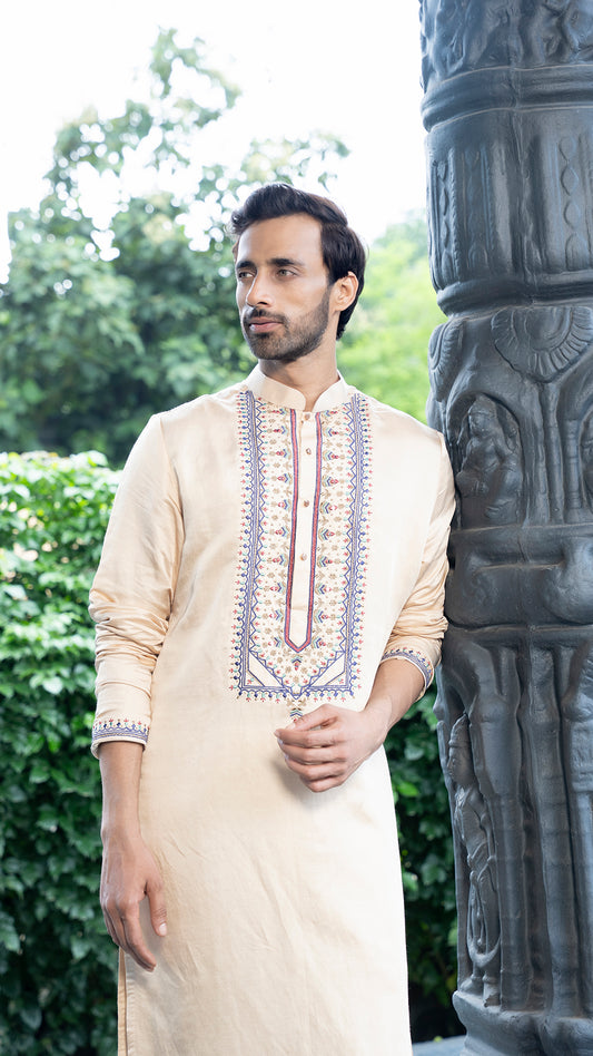 Man wearing a traditional beige kurta with embroidery standing next to an ornate stone column.