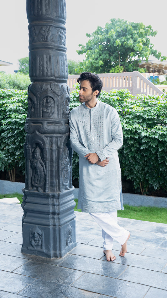 Man in traditional attire standing next to a stone pillar with carvings in an outdoor setting.