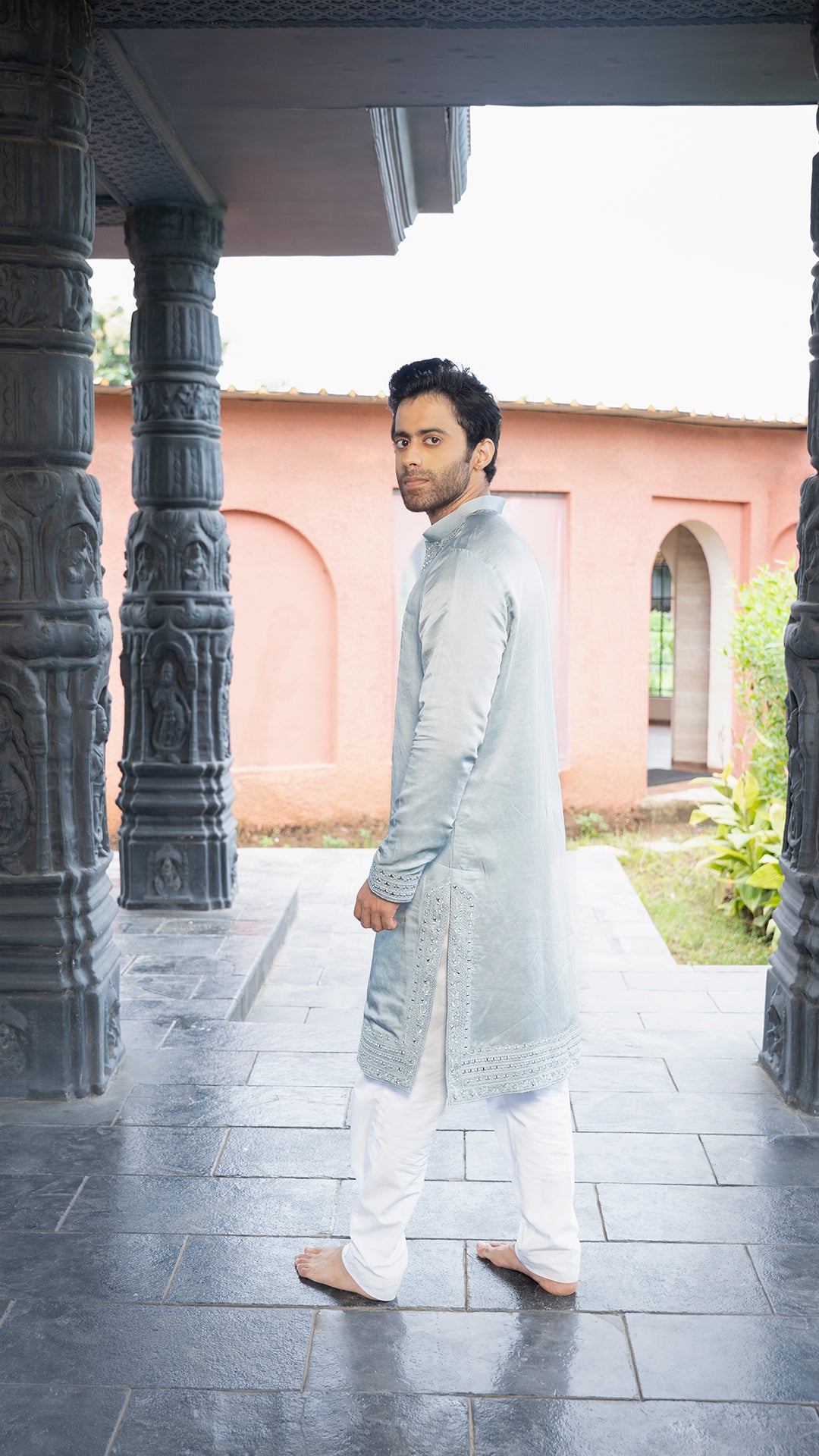 Man in traditional attire standing under a architectural structure with a pink building in the background.