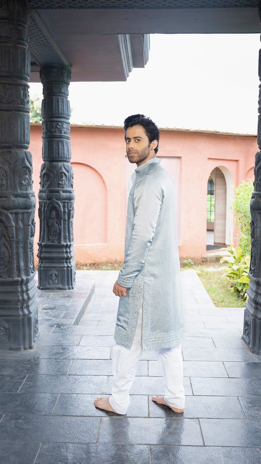 Man in traditional attire standing under a architectural structure with a pink building in the background.
