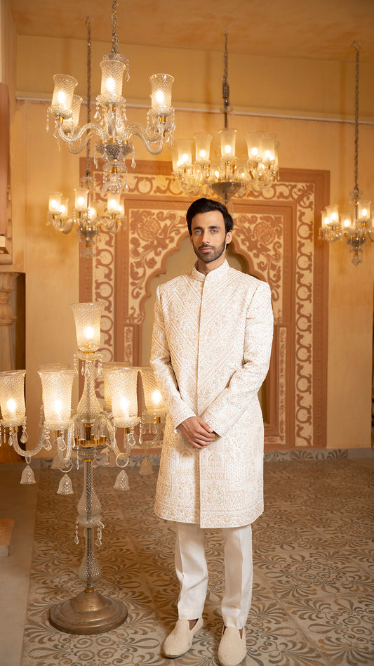 Man in traditional white sherwani standing in a decorated room with chandeliers.