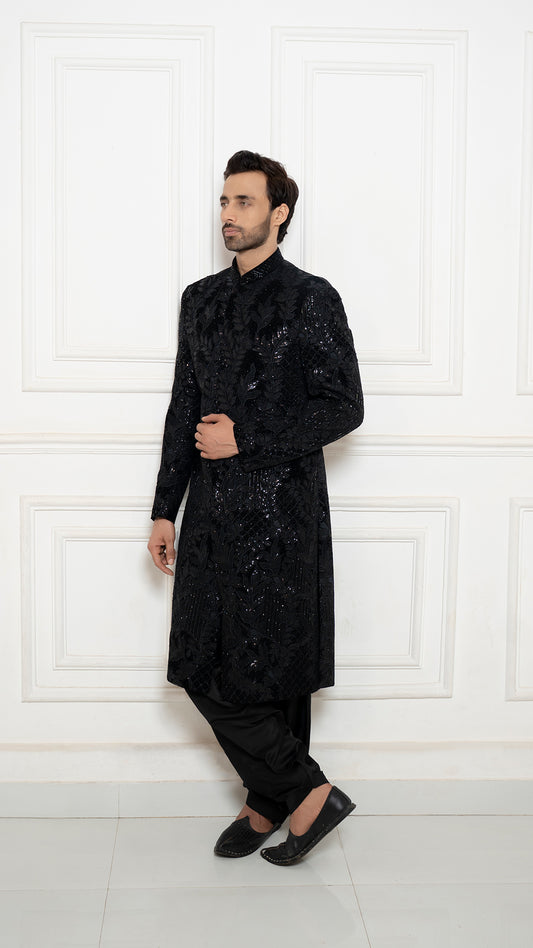Man wearing a black sequined kurta with black pants against a white wall.