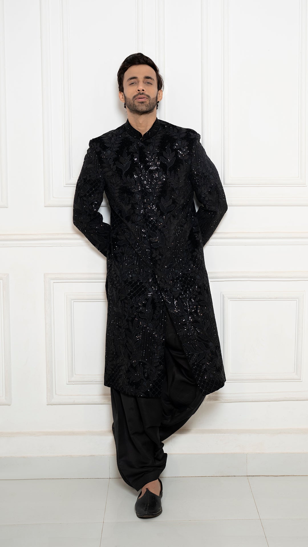 Man wearing a black, ornate traditional outfit against a white paneled wall.