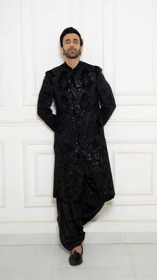 Man wearing a black, ornate traditional outfit against a white paneled wall.