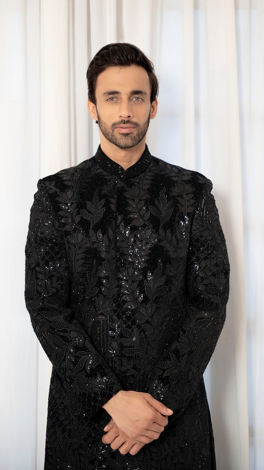 Man wearing a black embroidered sherwani against a white curtain background