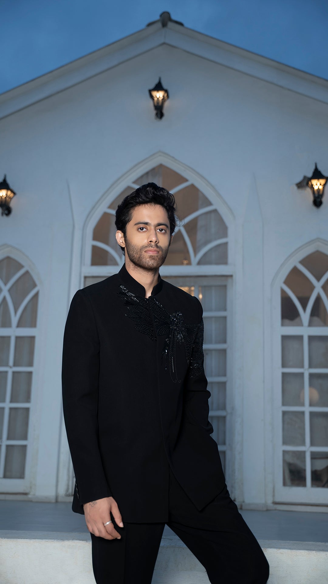 Man in a black jodhpuri standing in front of a white building with arched windows.