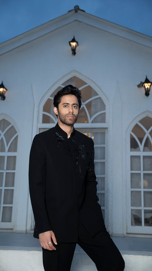 Man in a black jodhpuri standing in front of a white building with arched windows.