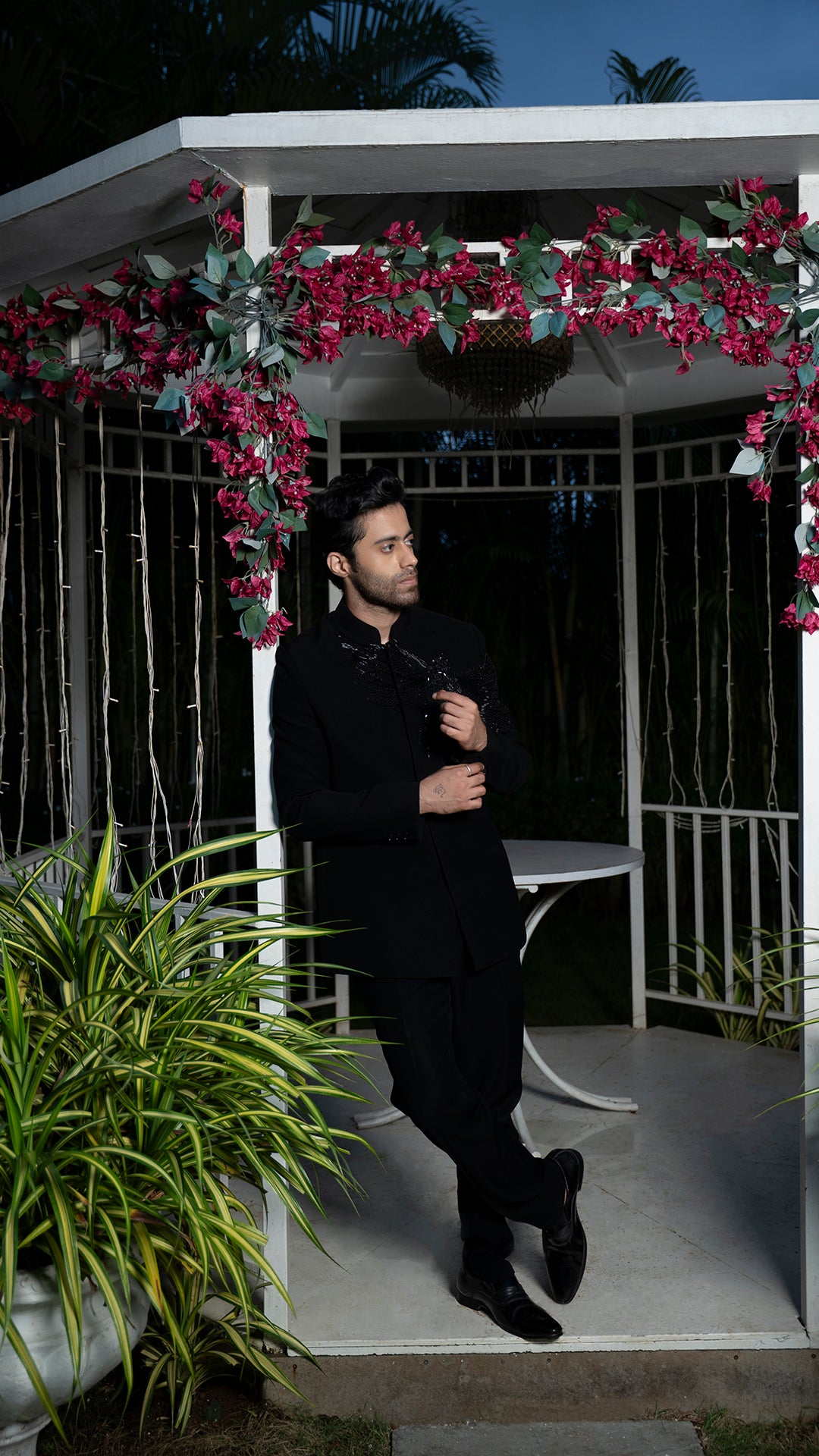 Man in a black jodhpuri suit standing under a floral archway at night.