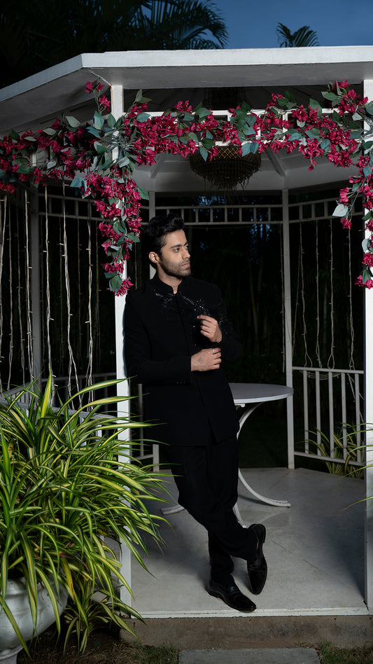 Man in a black jodhpuri suit standing under a floral archway at night.