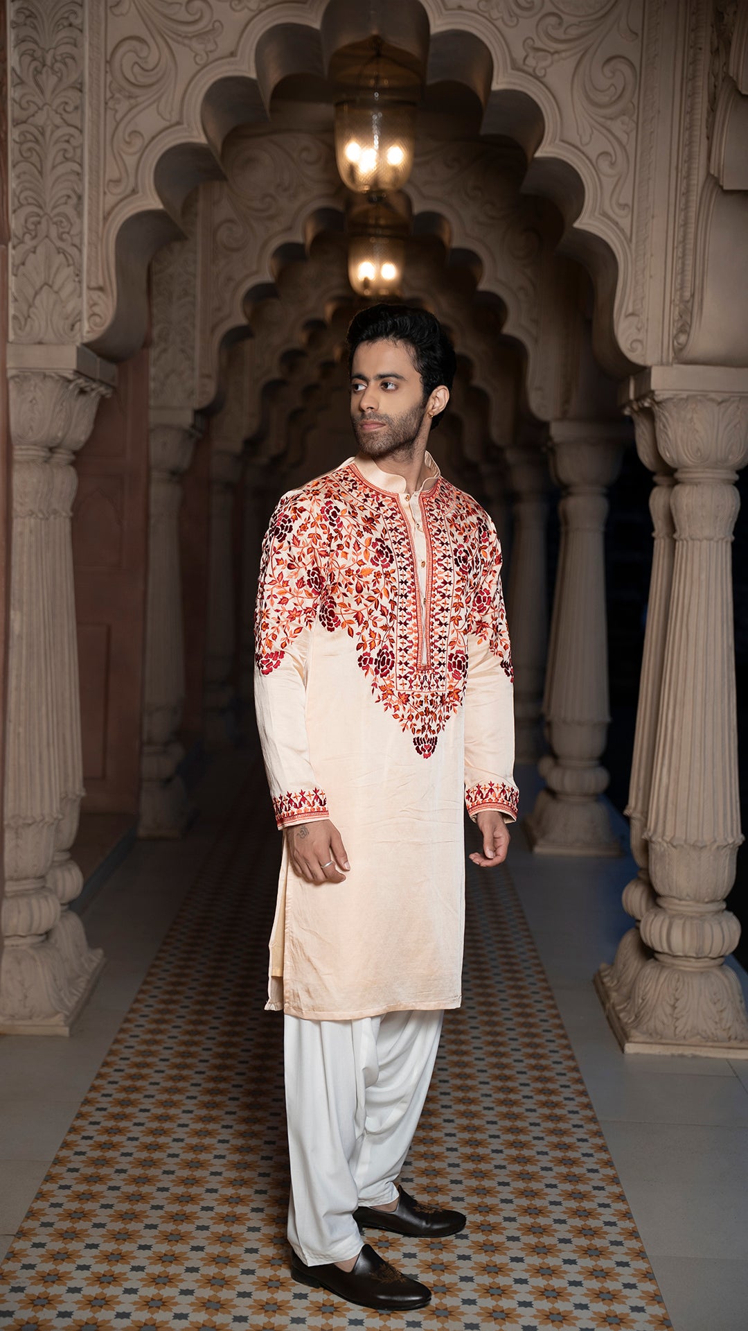 Man wearing a traditional cream kurta with red embroidery in an ornate architectural setting.