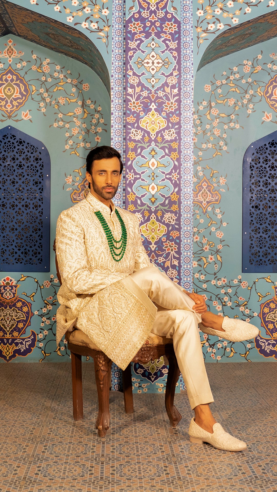 Man in traditional attire sitting on a chair against a decorative wall.